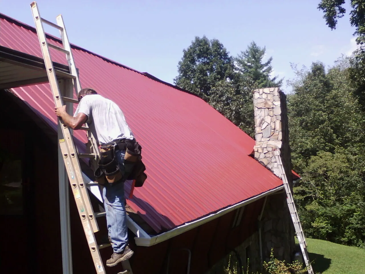 Roofer on a ladder beside a red metal roof and stone chimney — Soffit & Fascia Repair by Roofing Brothers in Latham